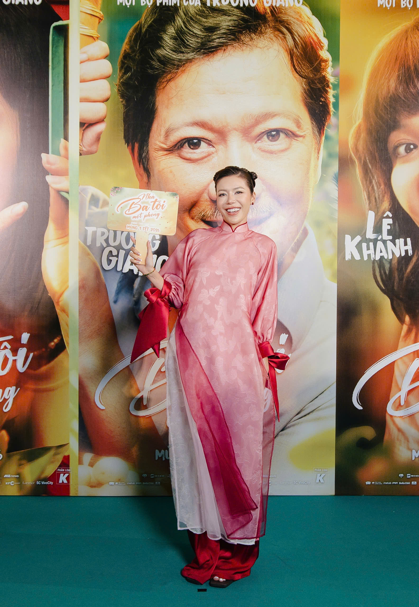 Minh Tú diện áo dài cách tân gam hồng phấn, chất liệu voan mỏng nhẹ được xử lý layer tinh tế, tạo hiệu ứng chuyển sắc mềm mại và bay bổng. Phần tay áo phồng vừa phải, nhấn bằng nơ đỏ đậm ở cổ tay mang lại điểm nhấn trẻ trung, trong khi quần lụa đỏ ton-sur-ton giúp tổng thể hài hòa và nổi bật. Kiểu tóc búi gọn gàng cùng lối trang điểm tươi tắn càng tôn lên nụ cười rạng rỡ của nàng mẫu.