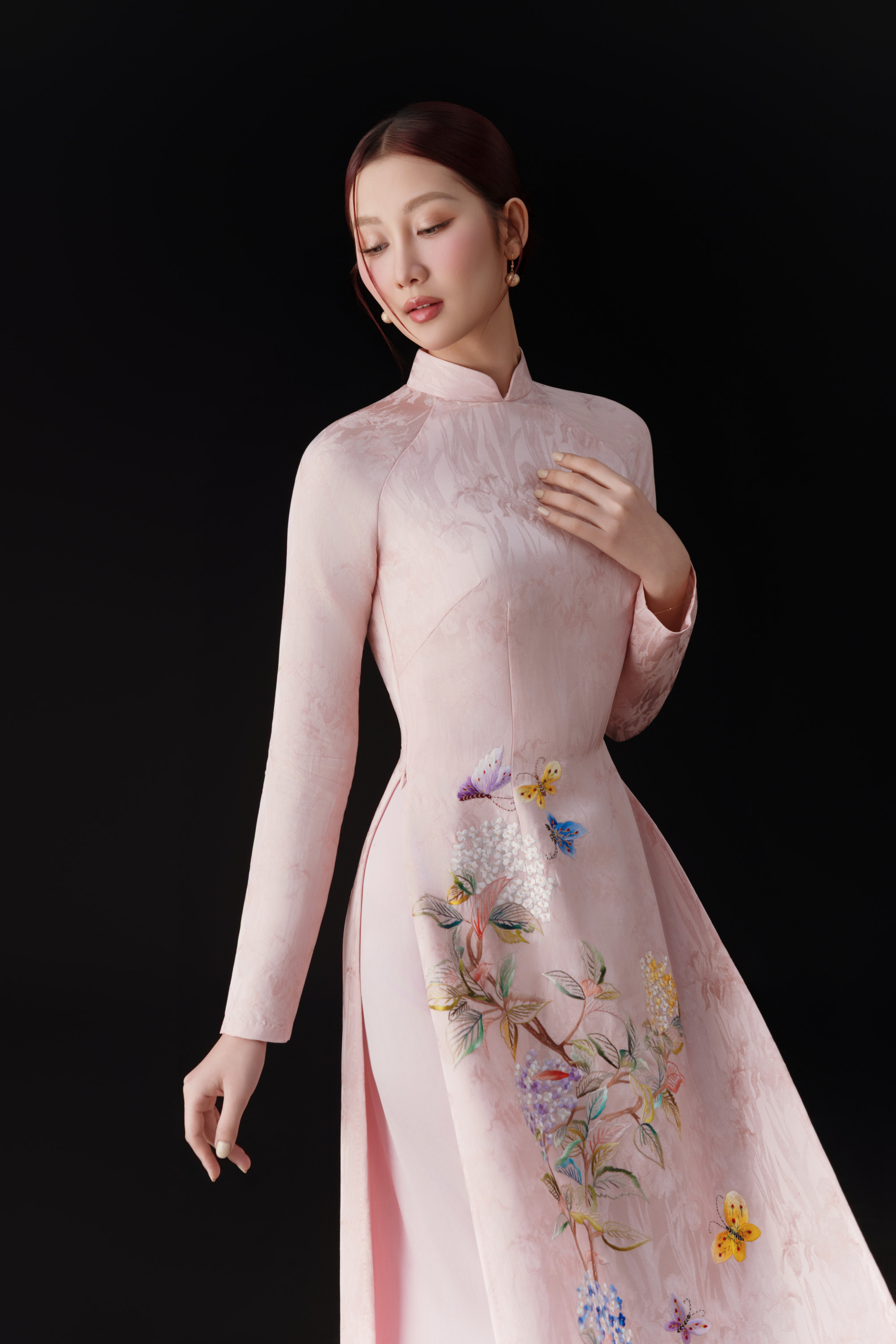 Quế Anh toát lên vẻ đẹp dịu dàng và tinh khôi khi diện áo dài lụa màu hồng pastel ôm dáng vừa vặn, kết hợp họa tiết thêu hoa lá và bướm tinh xảo, càng làm nổi bật vóc dáng thanh mảnh và nét mềm mại. 