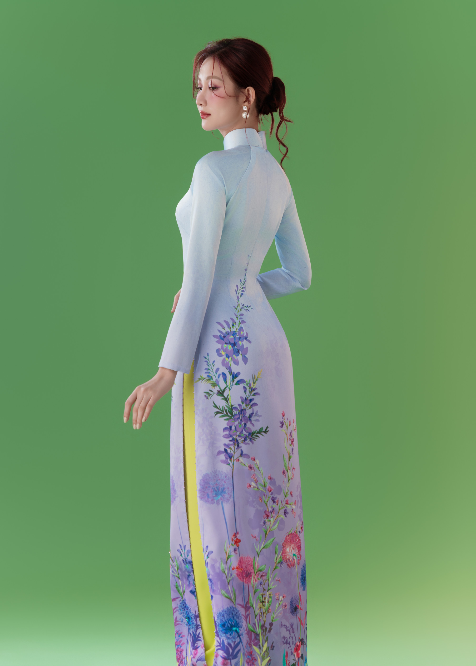 Mẫu áo dài gam xanh pastel chuyển sắc tím nhạt, in họa tiết hoa cỏ mềm mại, ôm khéo dáng người, giúp Quế Anh trở nên thanh lịch và quyến rũ một cách tinh tế. Kiểu trang điểm trong veo với nền mỏng tự nhiên, má hồng phớt nhẹ, đôi môi hồng đào căng bóng càng giúp nàng hậu thêm cuốn hút. Tóc búi thấp gọn gàng, điểm vài lọn tóc uốn rủ sau gáy, kết hợp khuyên tai ngọc trai tạo cảm giác cổ điển, trang nhã.
