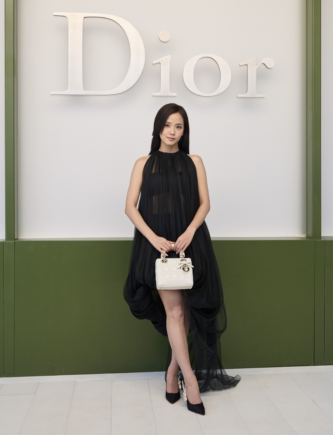 Jisoo ghi điểm với hình ảnh thanh lịch pha nét hiện đại rất đặc trưng. Cô chọn thiết kế váy đen dáng suông bằng chất liệu voan mỏng, tạo hiệu ứng mềm mại và bay bổng theo từng bước di chuyển. Phom váy tối giản nhưng được xử lý khéo léo ở phần độ dài bất đối xứng, giúp tổng thể vừa cổ điển vừa thời thượng. Jisoo hoàn thiện diện mạo bằng túi xách Lady Dior màu trắng và giày cao gót mũi nhọn ton-sur-ton, tạo điểm nhấn tinh tế trên nền trang phục tối màu. 