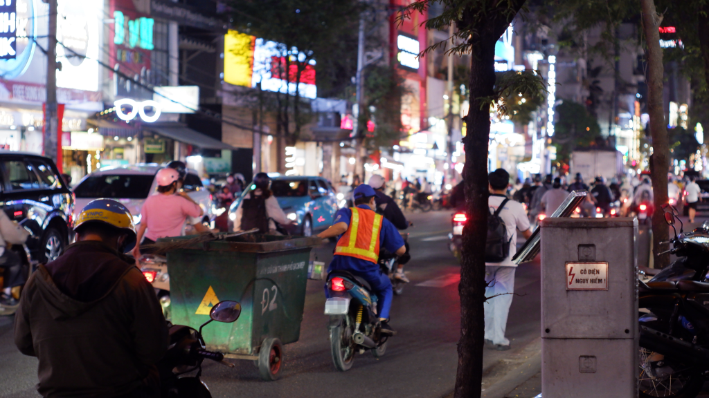 Công nhân vệ sinh môi trường lái xe máy bằng một tay, tay còn lại kéo theo xe thu gom rác trên đường Nguyễn Gia Trí. Ảnh: Huy Trình.