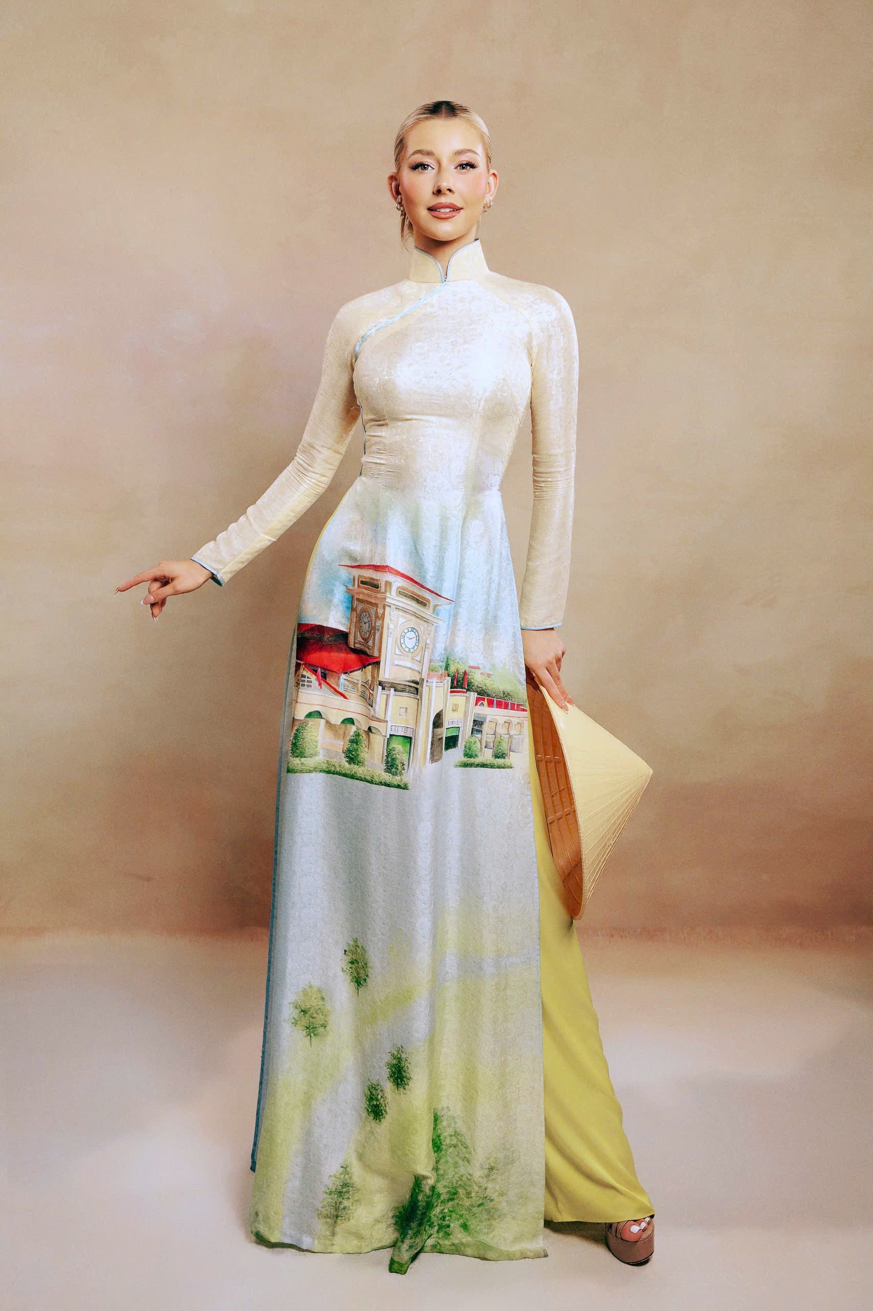 Trong đó, với BST Thành phố vươn mình, anh tái hiện những công trình, cảnh sắc nổi bật của thành phố. Trong ảnh, Miss Cosmo 2025 - Hoa hậu Hoàn vũ Quốc tế 2025 Yolina Lindquist diện áo dài với tông vàng nhạt làm chủ đạo, thể hiện quang cảnh chợ Bến Thành, công trình có tuổi đời hơn 100 năm, là điểm đến nổi tiếng của TPHCM nhiều năm qua.