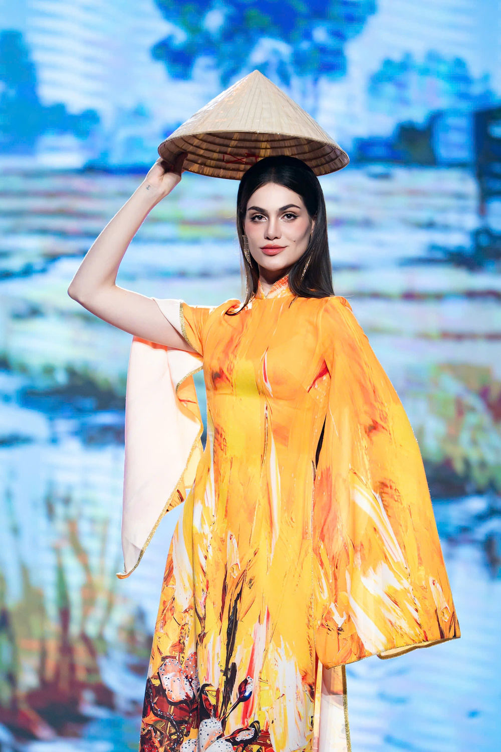 Miss Charm 2025 Anna Blanco xuất hiện đầy cuốn hút với phong cách thời trang tôn vinh vẻ đẹp văn hóa Việt Nam. Cô diện áo dài màu vàng cam rực rỡ với phom dáng ôm sát, tôn lên đường cong thanh thoát và thần thái tự tin trên sân khấu. Thiết kế gây ấn tượng bởi những mảng màu chuyển sắc như nét cọ hội họa, kết hợp họa tiết phong cảnh đồng quê gợi liên tưởng đến vẻ đẹp yên bình của làng quê Việt.