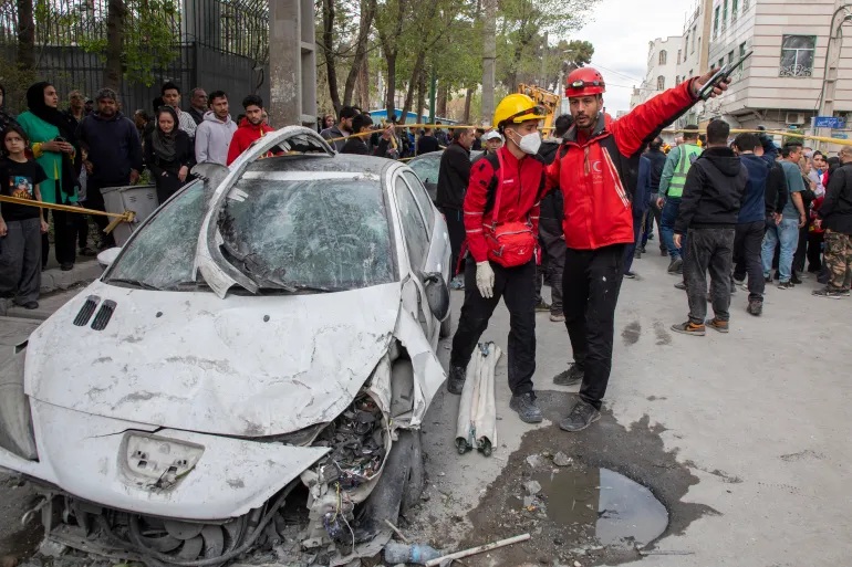 Các nhân viên cứu hộ và người dân đứng gần những chiếc xe và một tòa nhà dân cư bị trúng bom trong cuộc không kích vào rạng sáng ngày 27/3, tại Tehran, Iran - Ảnh: Majid Saeedi/Getty Images