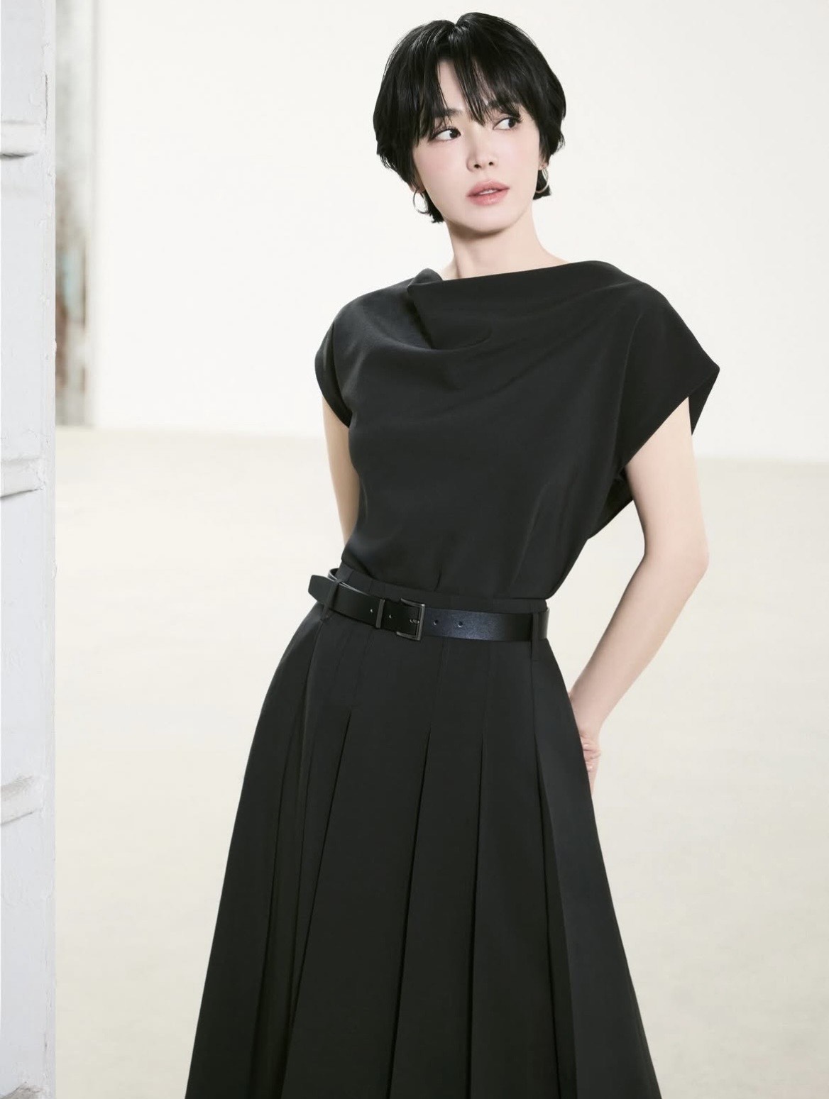 Chiếc đầm đen dáng dài, thắt eo bằng belt là lựa chọn hoàn hảo cho những ngày cần sự chỉn chu tuyệt đối. Phom dáng gọn gàng, không chi tiết thừa giúp tôn lên thần thái tự tin. Với outfit tối giản, Song Hye Kyo chọn kiểu trang điểm có phần sắc sảo hơn, với eyeliner mảnh và son tông đất, mang đến hình ảnh người phụ nữ công sở hiện đại, độc lập.
