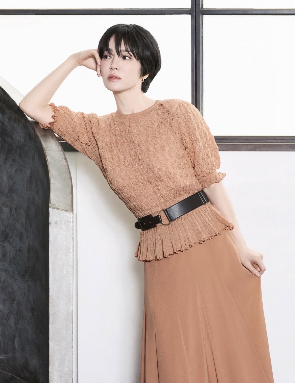 Nữ diễn viên lựa chọn áo dệt kim tay ngắn phối cùng chân váy nâu dáng dài. Thiết kế nhún nhẹ ở eo và tay áo tạo cảm giác mềm mại, nữ tính. Makeup vẫn trung thành với phong cách tự nhiên: làn da căng bóng, má ửng nhẹ và môi tông cam đất, rất phù hợp với không khí mùa hè.