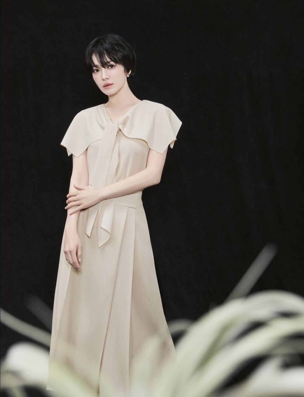 Diện chiếc váy dài màu be với thiết kế mềm mại, điểm nhấn là phần cổ thắt nơ nhẹ nhàng và tay áo rủ, Song Hye Kyo mang đến cảm giác bay bổng, nữ tính. Gam màu trung tính không chỉ tôn lên làn da mà còn mang lại vẻ trang nhã, tinh khôi. Phom dáng váy suông dài giúp tổng thể trở nên thanh thoát, kín đáo nhưng không hề đơn điệu. Kiểu tóc ngắn gọn gàng kết hợp cùng lối trang điểm tự nhiên càng làm nổi bật thần thái nhẹ nhàng, sang trọng của ngôi sao hàng đầu xứ Hàn.