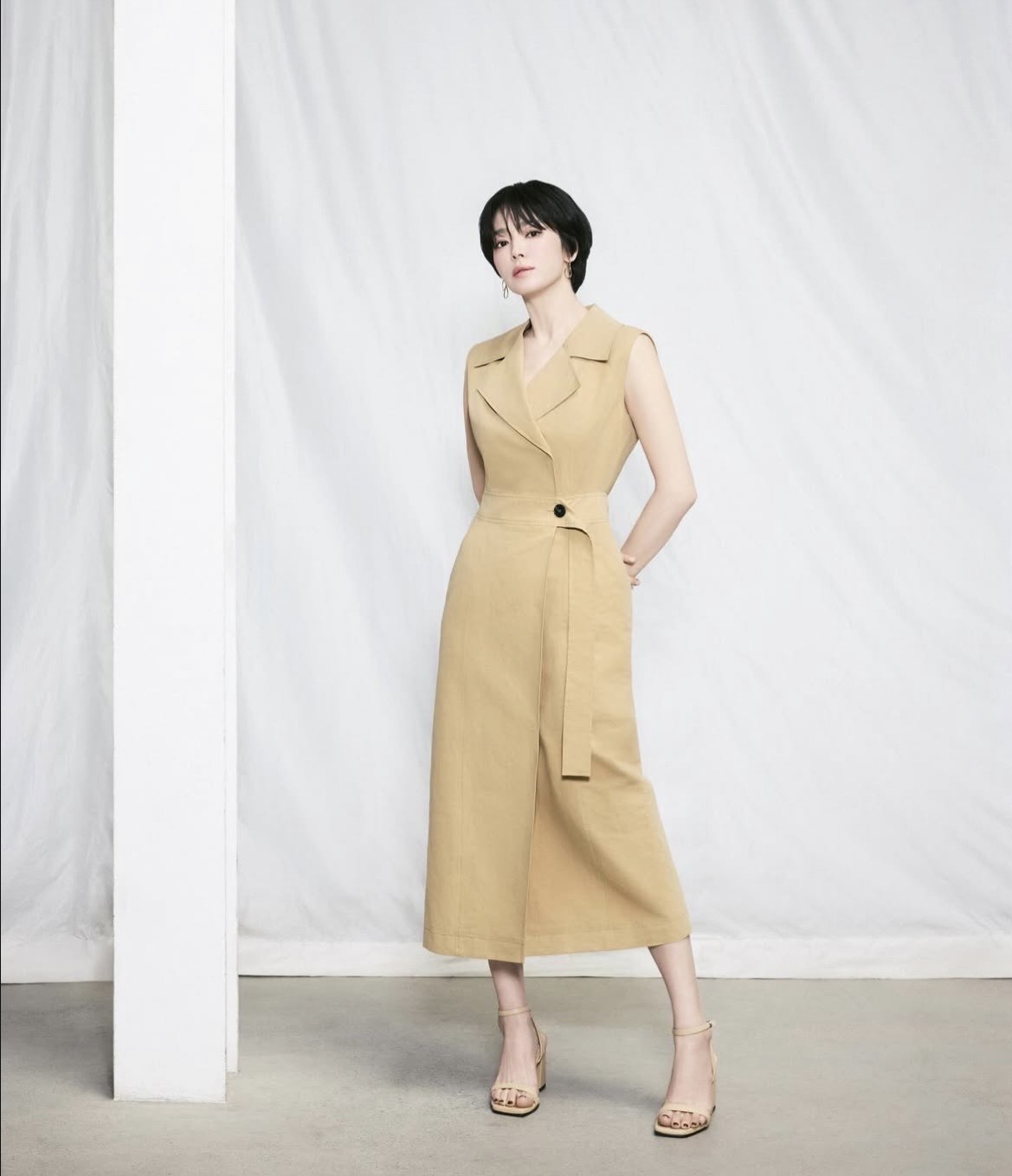 Thiết kế váy liền thân không tay với phom dáng chuẩn chỉnh, lấy cảm hứng từ kiểu vest, tạo nên vẻ ngoài vừa mạnh mẽ vừa tinh tế cho Song Hye Kyo. Phần cổ áo ve và chi tiết thắt eo giúp tôn lên vóc dáng gọn gàng, đồng thời mang lại cảm giác chuyên nghiệp, thời thượng. Gam màu vàng be ấm áp kết hợp cùng đôi sandal cao gót thanh mảnh tô điểm cho trang phục càng thêm phần thanh thoát và nữ tính. 