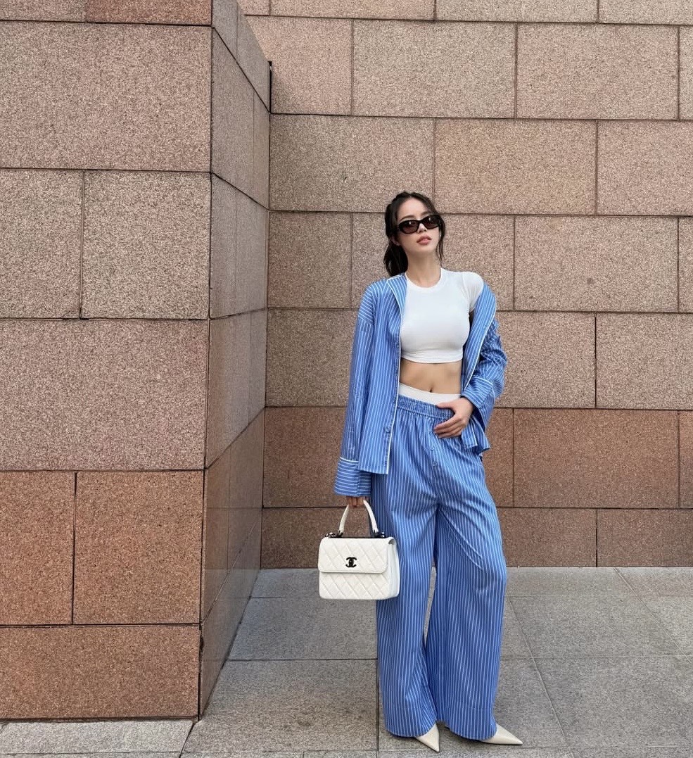 Hoa hậu Thanh Thuỷ đến hình ảnh mùa hè hiện đại, phóng khoáng nhưng vẫn giữ được nét thanh lịch đặc trưng. Cô lựa chọn set đồ tông xanh sọc gồm áo sơ mi khoác ngoài và quần ống rộng đồng điệu, kết hợp cùng áo croptop trắng ôm sát, khoe khéo vòng eo thon gọn. Tổng thể trang phục vừa thoải mái, vừa pha chút tinh thần menswear cá tính. Điểm nhấn nằm ở chiếc túi xách trắng cổ điển và giày mũi nhọn ton-sur-ton, giúp cân bằng giữa sự năng động và nét sang trọng. 
