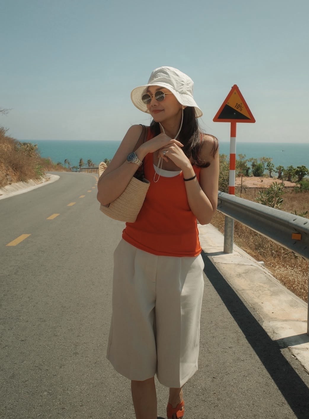 Giữa khung cảnh biển xanh nắng gắt, phong cách mùa hè của Thanh Hằng gây ấn tượng trước hết bởi tinh thần phóng khoáng và trẻ trung. Không chọn những thiết kế cầu kỳ, cô ưu tiên áo tank top sắc cam nổi bật phối cùng quần bermuda dáng rộng tông trung tính, tạo nên sự cân bằng giữa năng động và thanh lịch. Điểm nhấn nằm ở loạt phụ kiện mang hơi hướng du lịch như mũ bucket trắng, kính râm tròn và túi cói, vừa tăng tính thời trang vừa đảm bảo sự tiện dụng dưới nắng hè. Tổng thể trang phục mang đến cảm giác thoải mái, dễ di chuyển nhưng vẫn giữ được thần thái thời thượng đặc trưng. 