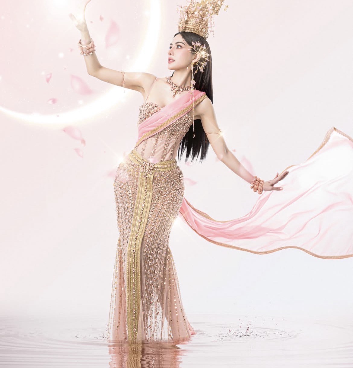 Trong bộ ảnh mừng lễ hội Songkran, Miss Grand Thái Lan 2026 Ning Patthama ghi điểm với hình ảnh tựa nữ thần truyền thống Thái Lan bước ra từ huyền thoại. Người đẹp lựa chọn trang phục mang đậm tinh thần văn hóa bản địa với phom dáng ôm sát, được đính kết cầu kỳ bằng các chi tiết ánh kim và pha lê, tôn lên đường cong mềm mại. Dải lụa hồng vắt chéo qua vai, bay bổng theo từng chuyển động, tạo hiệu ứng thị giác uyển chuyển như dòng nước - biểu tượng đặc trưng của Songkran.