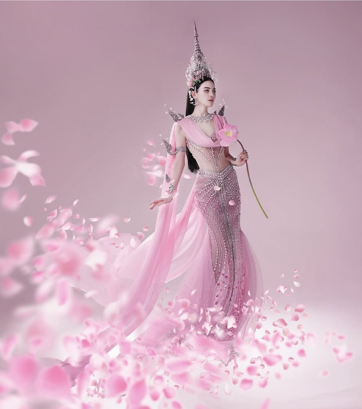 Trong bộ ảnh mới, Mai Davika khoác lên mình trang phục mang cảm hứng truyền thống Thái Lan với tông hồng chủ đạo, sắc màu tượng trưng cho sự dịu dàng và may mắn. Bộ váy được thiết kế ôm sát cơ thể, tôn lên vóc dáng thanh mảnh, kết hợp cùng kỹ thuật đính kết tinh xảo với họa tiết hoa văn truyền thống.    Phần thân trên được xử lý bằng chất liệu xuyên thấu nhẹ, tạo hiệu ứng mềm mại nhưng vẫn sang trọng. Dải lụa vắt chéo qua vai gợi nhớ đến trang phục cổ điển của các vũ nữ cung đình Thái, mang lại cảm giác bay bổng, uyển chuyển như bước ra từ thần thoại.