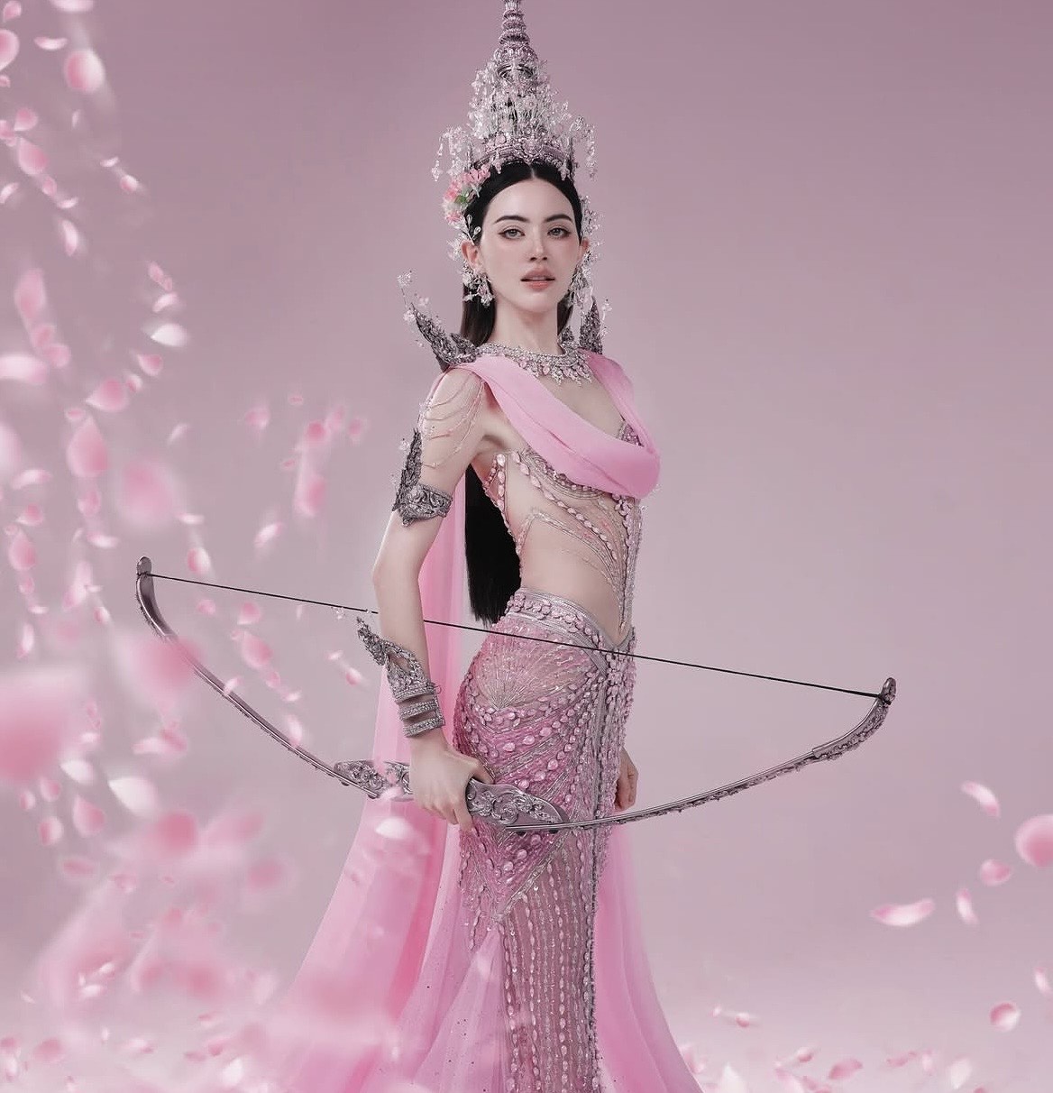 Điểm nhấn nổi bật chính là chiếc mũ đội đầu cao vút, được chế tác cầu kỳ với các chi tiết kim loại và pha lê lấp lánh - biểu tượng thường thấy trong tạo hình các nữ thần hoặc nhân vật hoàng tộc trong văn hóa Thái Lan. Đi kèm là bộ trang sức đồng bộ gồm vòng cổ, hoa tai và vòng tay bản lớn, góp phần hoàn thiện diện mạo quyền quý.