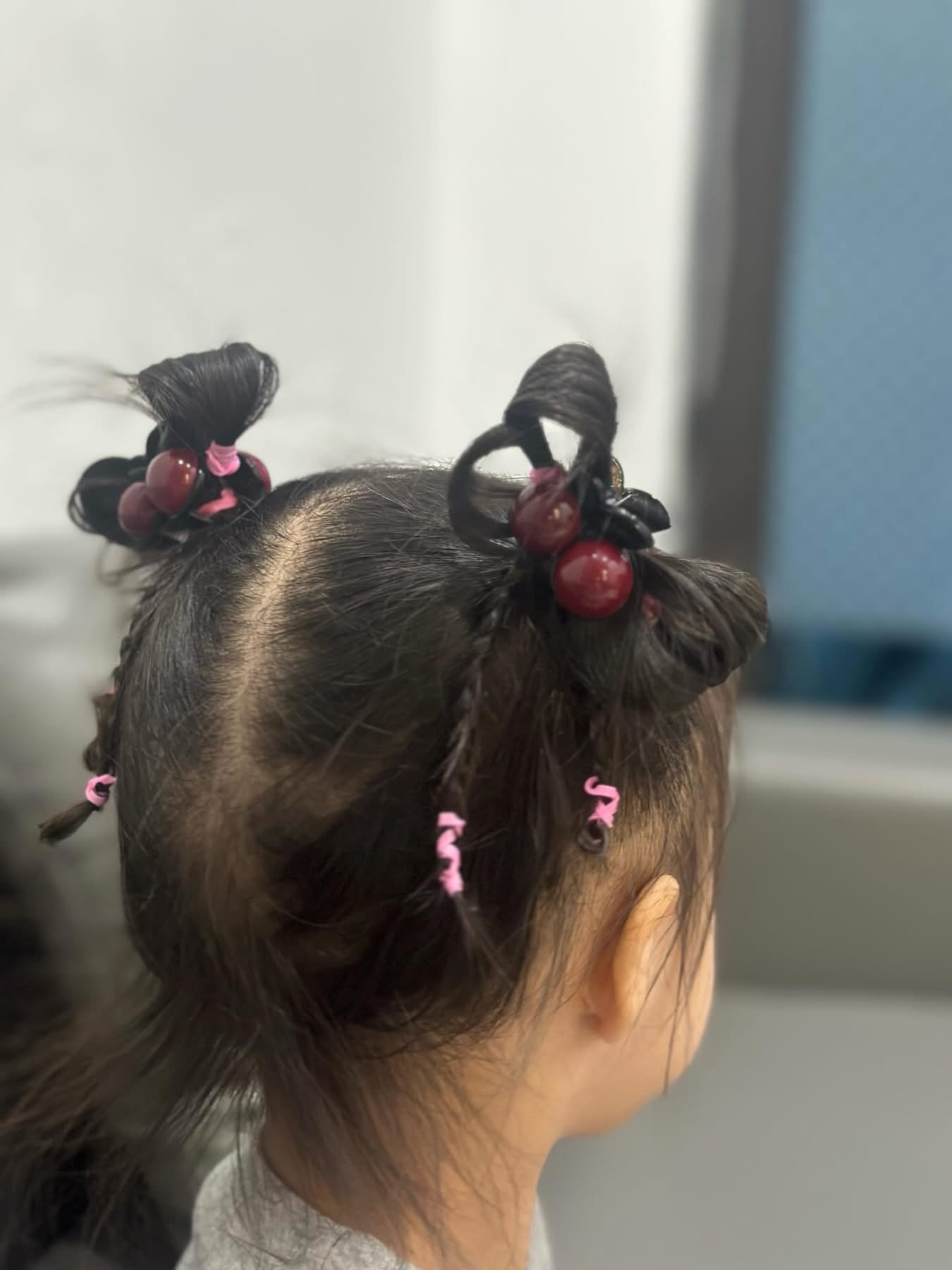 Kiểu tóc này đúng chuẩn 'vườn trái cây di động'. Hai bên đầu bé được búi gọn thành hai chỏm xinh xắn, rồi khéo léo 'cài cắm' thêm những quả nhỏ đỏ mọng nhìn cứ như… chùm nho lạc vào mái tóc. Phía dưới còn có vài lọn tóc tết bé xíu, buộc thun hồng xoăn tạo kiểu độc đáo. Cách làm thì nghe có vẻ cầu kỳ nhưng thật ra cũng chỉ cần chút kiên nhẫn. Trước hết chia tóc làm 2 phần đều nhau, buộc cao lên rồi xoắn lại thành búi nhỏ. Tiếp theo, lấy phụ kiện gài quanh búi cho thêm phần nổi bật. Phần tóc còn lại chia nhỏ, tết vài bím mini rồi buộc thun cho sinh động.