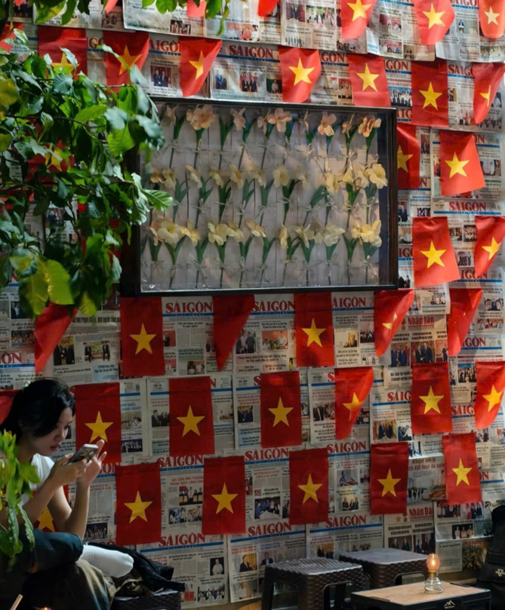 Bước vào bên trong, quán tiếp tục khai thác mạnh yếu tố hoài niệm với thiết kế mở, tận dụng ánh sáng tự nhiên để làm nổi bật các chi tiết trang trí. Nổi bật nhất là góc check-in với bức tường được phủ kín bằng những tờ báo cũ – gợi nhớ đến dòng chảy lịch sử và ký ức một thời – xen kẽ là hình ảnh lá cờ đỏ sao vàng được bố trí có chủ đích, tạo nên một không gian vừa mang tính nghệ thuật vừa giàu chiều sâu văn hóa.