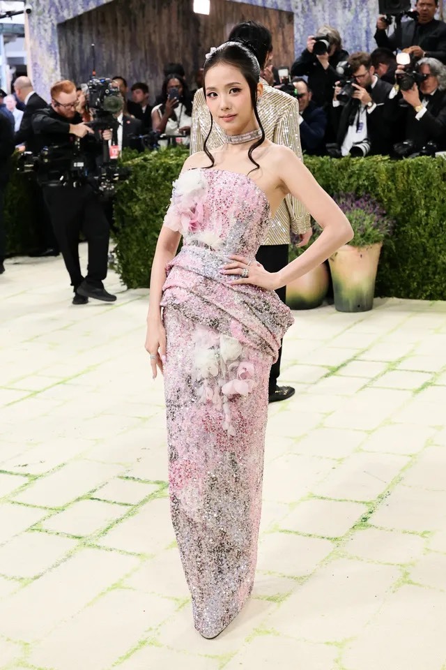 Xuất hiện tại Met Gala, Jisoo ghi dấu ấn với phong cách thanh lịch đặc trưng của Dior với chiếc đầm dạ hội tông hồng pastel ôm dáng, phủ sequin lấp lánh nhẹ nhàng như lớp sương, mang đến cảm giác nữ tính nhưng không kém phần sang trọng. Thiết kế lệch vai kết hợp chi tiết draping mềm mại và những điểm nhấn hoa 3D tạo chiều sâu thị giác, tôn lên vóc dáng thanh mảnh của cô một cách tinh tế. Kiểu tóc búi cao gọn gàng cùng những lọn tóc buông nhẹ giúp tổng thể thêm phần yêu kiều. Jisoo không chọn sự phô trương, mà chinh phục thảm đỏ bằng khí chất dịu dàng, chuẩn mực của một 'nàng thơ Dior' đích thực.