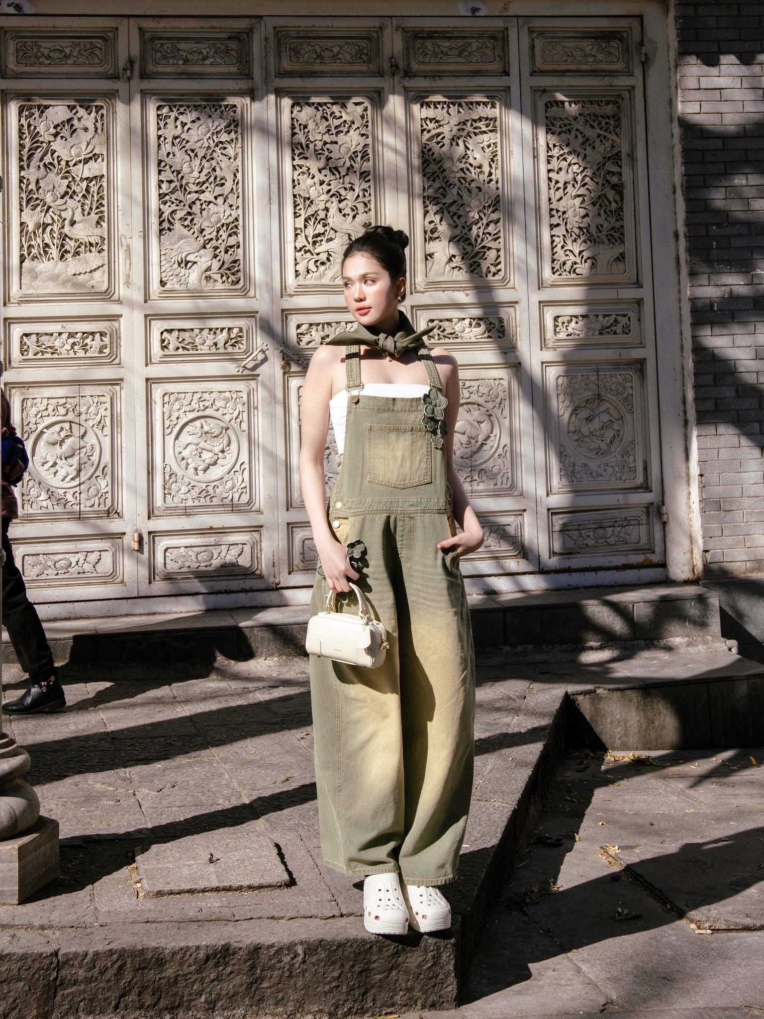 Bộ jumpsuit yếm dáng rộng xanh denim bạc màu với thiết kế suông thoải mái, tạo cảm giác vừa phóng khoáng vừa thời thượng. Bên trong là chiếc áo quây trắng giúp tổng thể trở nên nhẹ nhàng và nữ tính hơn. Điểm nhấn đặc biệt nằm ở chiếc khăn lụa buộc cổ, mang hơi hướng vintage châu Âu, giúp outfit thêm tinh tế và sang trọng. Ngọc Trinh kết hợp cùng túi xách mini màu kem và đôi giày trắng platform, tạo nên sự hài hòa giữa nét hiện đại và cổ điển.