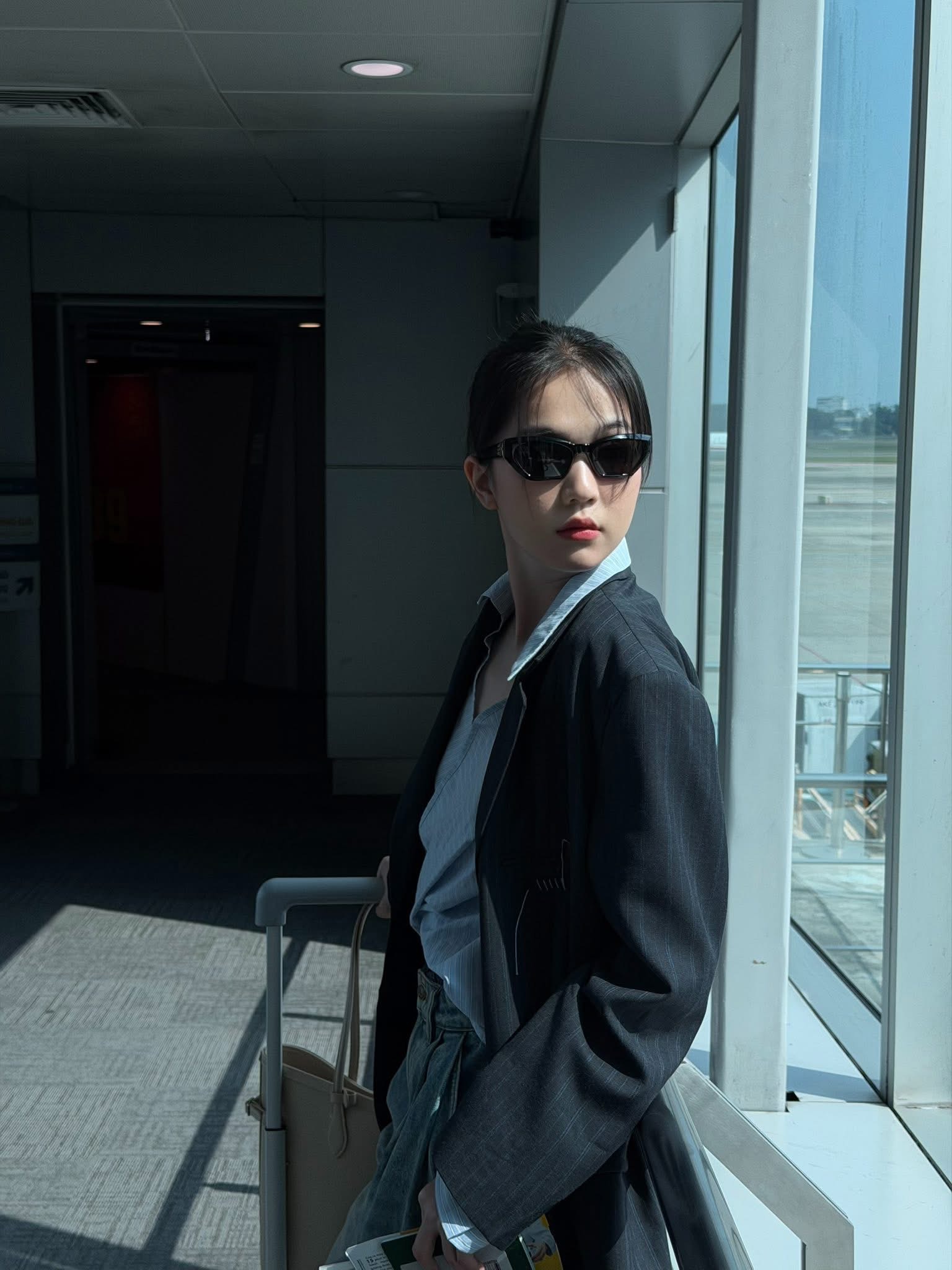 Ngọc Trinh toát lên vẻ thanh lịch, hiện đại và đầy khí chất “cool girl” thời thượng. Cô lựa chọn outfit mang tông màu trung tính với blazer oversized phối cùng áo sơ mi sáng màu, tạo nên hình ảnh tối giản nhưng cực kỳ sang trọng. Chiếc quần cạp cao giúp tổng thể thêm gọn gàng và tôn lên vóc dáng thanh mảnh. Kính râm đen là điểm nhấn đắt giá, mang lại thần thái bí ẩn, tự tin như một fashionista tại sân bay. Cách búi tóc gọn gàng cùng lối trang điểm nhẹ nhàng càng làm nổi bật phong cách chic hiện đại, vừa cá tính vừa tinh tế. Tổng thể outfit mang đậm cảm hứng “airport fashion” – đơn giản nhưng vẫn đủ nổi bật và cuốn hút.