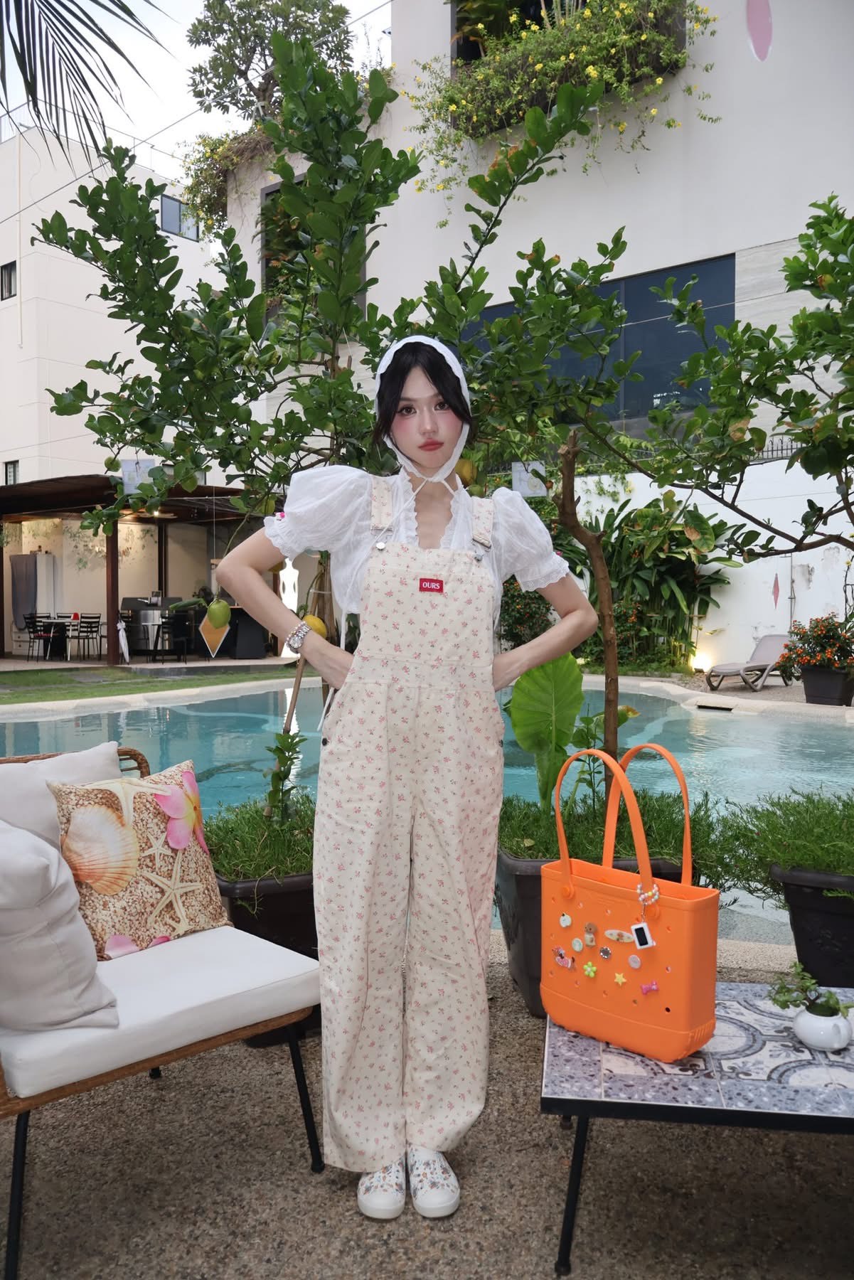 Người đẹp chọn jumpsuit hoa nhí dáng rộng phối áo blouse trắng tay phồng và khăn trùm đầu kiểu vintage. Đây là kiểu trang phục thiên về sự đáng yêu, kín đáo và mang cảm giác “cottagecore” nhẹ nhàng. Cách phối cùng túi màu cam nổi bật giúp tổng thể không bị quá bánh bèo mà vẫn giữ được nét trẻ trung, vui mắt.