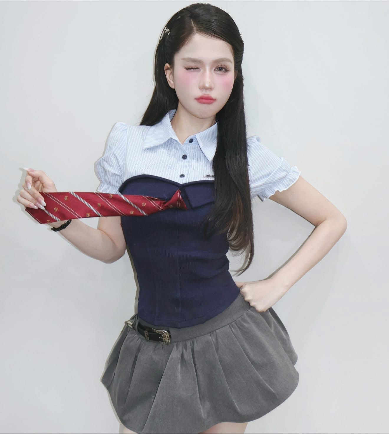 Ngọc Trinh diện áo corset phối sơ mi tay phồng cùng chân váy balloon màu xám. Tổng thể mang hơi hướng nữ sinh nhưng được xử lý theo phong cách thời trang hiện đại. Thay vì khoe vòng một hay đường cong quá đậm, outfit tập trung vào sự trẻ trung với phom dáng gọn gàng, gam màu navy – xám thanh lịch. Chiếc cà vạt đỏ tạo điểm nhấn tinh nghịch, giúp hình ảnh của Ngọc Trinh trông năng động hơn hẳn trước đây.