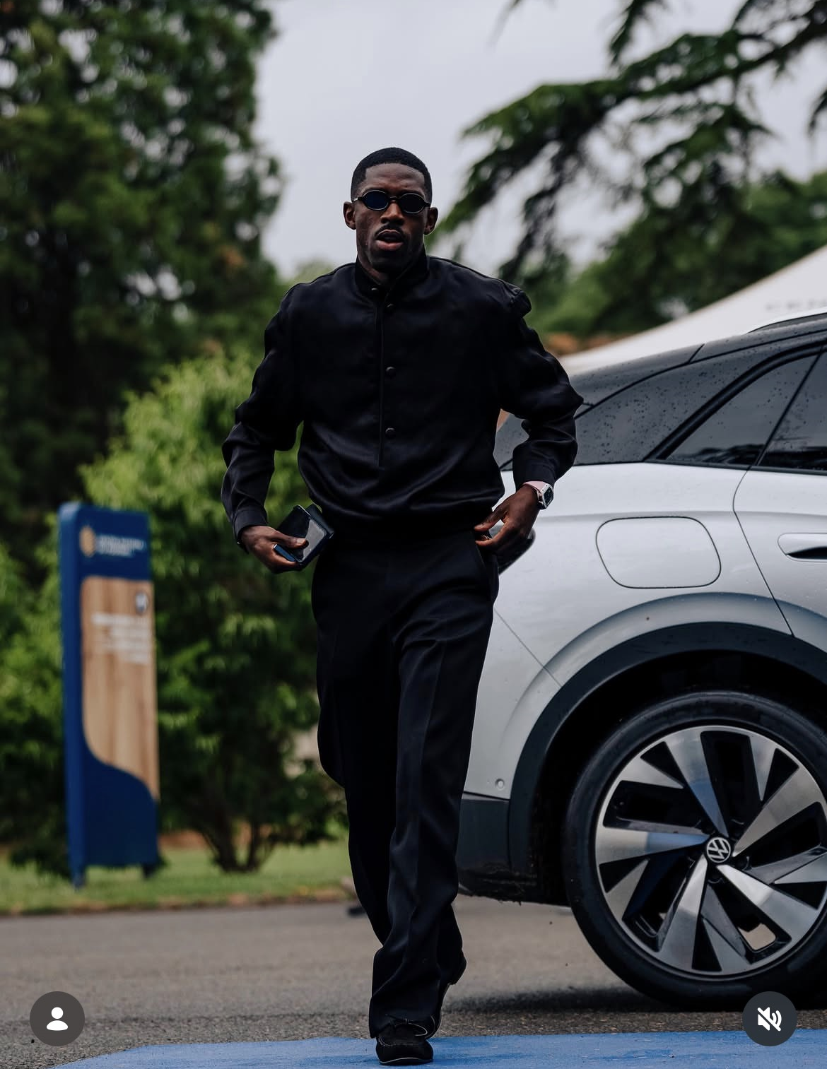 Ousmane Dembélé rũ bỏ hoàn toàn hình ảnh quần đùi số áo quen thuộc, Dembélé xuất hiện đầy quyền lực trong một set đồ all-black (nguyên cây đen) được thiết kế riêng. Anh diện chiếc áo khoác bomber cổ trụ cài cúc thanh lịch phối cùng quần tây đen ống đứng hoàn hảo. Điểm nhấn giúp nâng tầm bộ trang phục chính là những món phụ kiện kính râm đen gọng nhỏ cá tính, chiếc đồng hồ xa xỉ trên cổ tay và đôi giày da cao cấp. Outfit này có sự đồng hành thương hiệu cùng nhà mốt cao cấp Zegna, biến khoảnh khắc rời xe bước vào đại bản doanh của anh không khác gì một siêu mẫu đang chụp ảnh tạp chí.